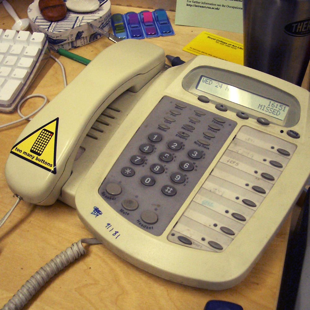 A 'Too many buttons' warning sticker, reading: our office phones have too many buttons, I don't know what most of them even do